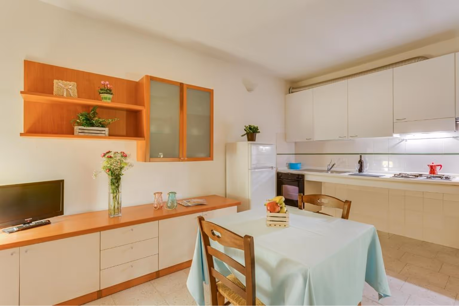 Світла кухня з бежевими шафами, столом із блакитною скатертиною та дерев'яними стільцями, холодильником і рослинами. Villaggio Mare Si