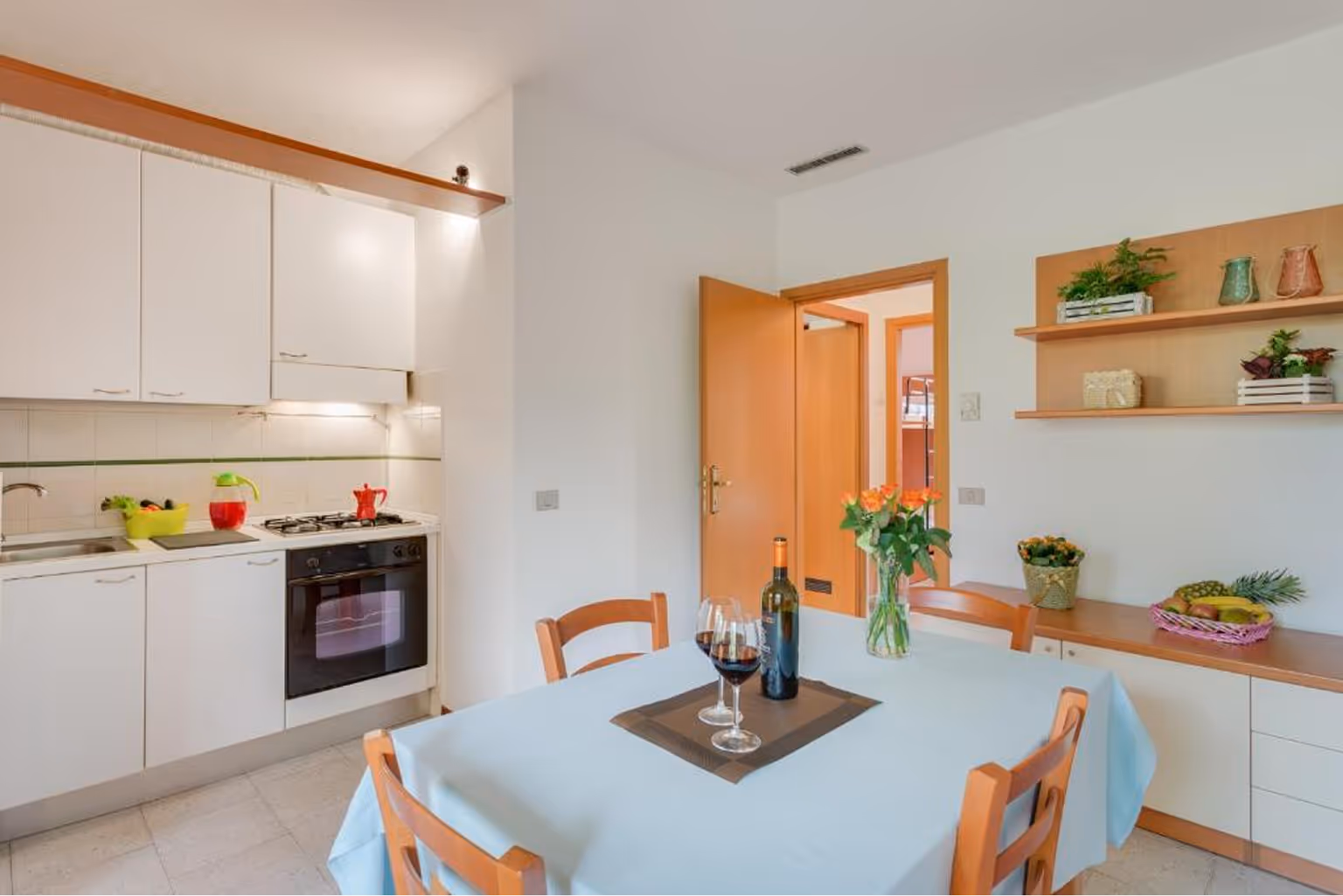 Світла кухня зі столом, накритим блакитною скатертиною, трьома дерев'яними стільцями, пляшкою вина і двома келихами з червоним вином. Villaggio Mare Si