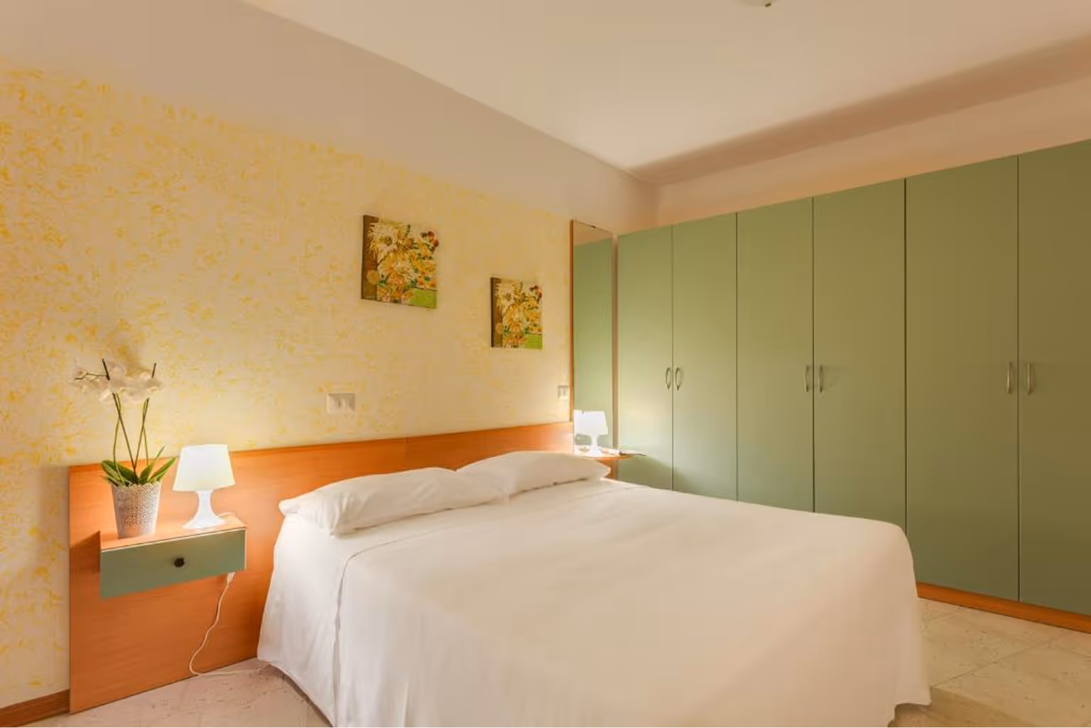 Світла спальня з двоспальним ліжком, зеленими шафами, нішею з лампою та квітковим горщиком на стіні з жовтим візерунком. Villaggio Mare Si