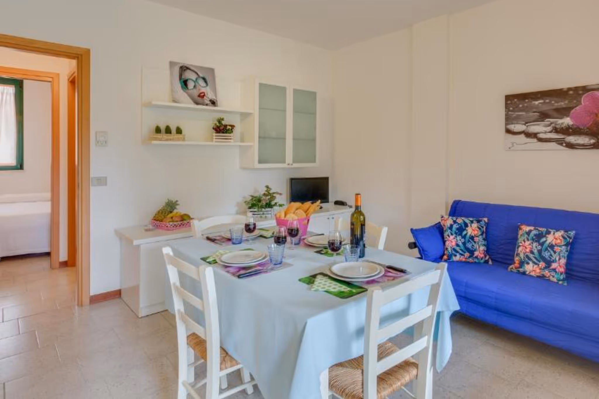 Їдальня з білим столом, накритим блакитною скатертиною, чотири стільці, пляшка вина, келихи з вином, корзина з хлібом та синій диван із квітковими подушками. Villaggio Mare Si