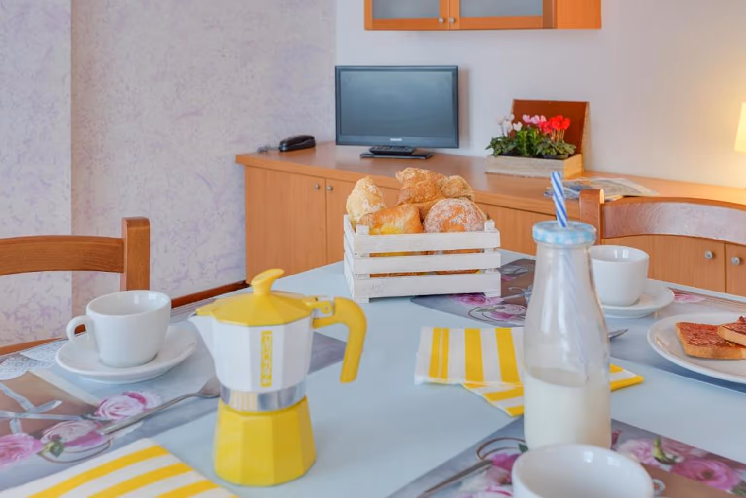 Сніданок на столі з жовтою кавовою кавоваркою, пляшкою молока, чашками, кошиком з хлібом і тостами, на фоні меблі та телевізор. Villaggio Mare Si