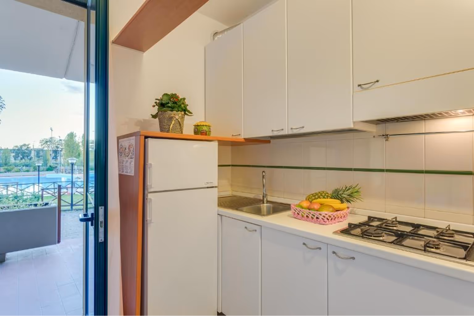 Світла кухня з білим холодильником, раковиною, варильною плитою і кошиком із фруктами на стільниці поруч із відкритими дверима на вулицю. Villaggio Mare Si