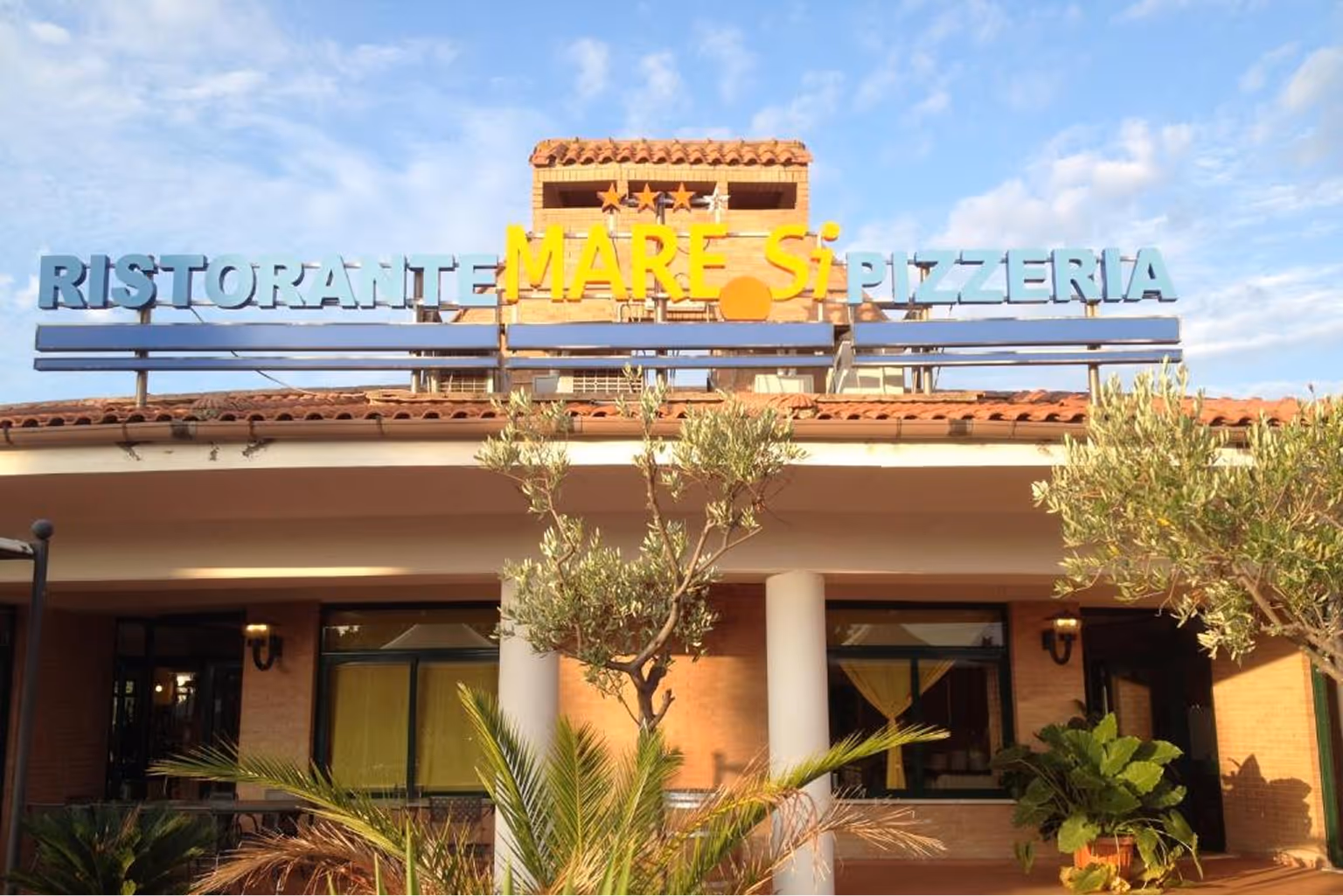 Фасад італійського ресторану з вивіскою Ristorante Mare Si Pizzeria на тлі блакитного неба. Villaggio Mare Si