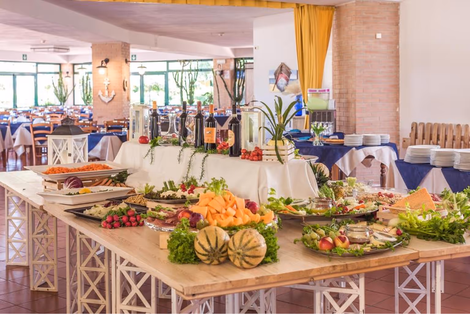 Стол із різноманітними стравами, фруктами, овочами та пляшками вина в яскраво освітленому ресторані з синіми накриттями на столах у фоні. Villaggio Mare Si