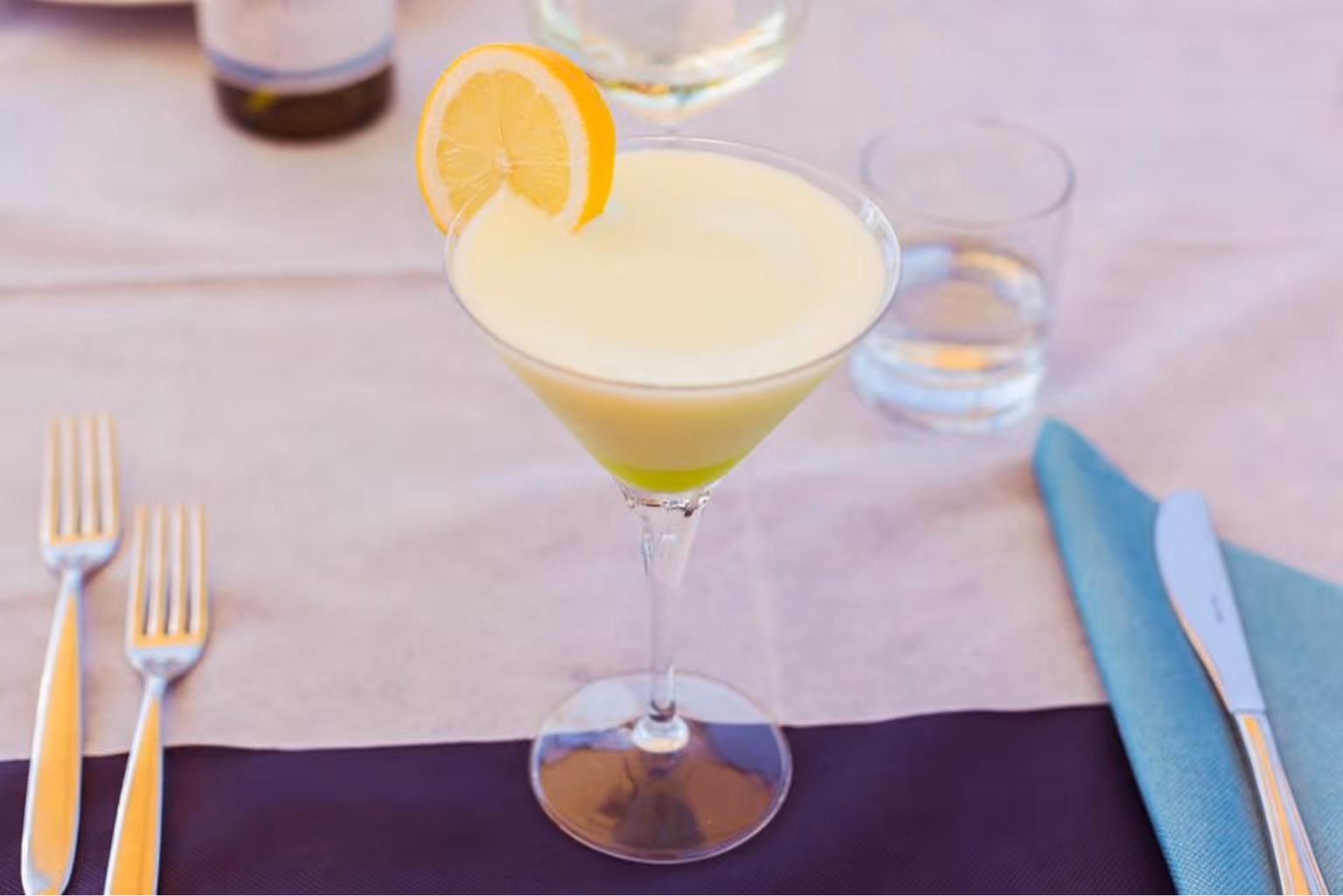 Коктейль з жовтою піною та скибочкою лимона на келиху, стоїть на столі з виделками, ножем і склянкою води. Villaggio Mare Si