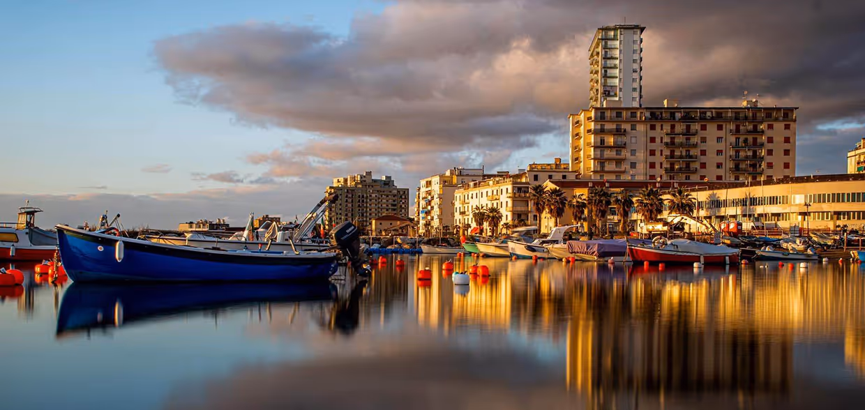 Маленькі човни на спокійній воді гавані з відбиттям будинків на тлі хмарного неба під час заходу сонця. Follonica, Toscana
