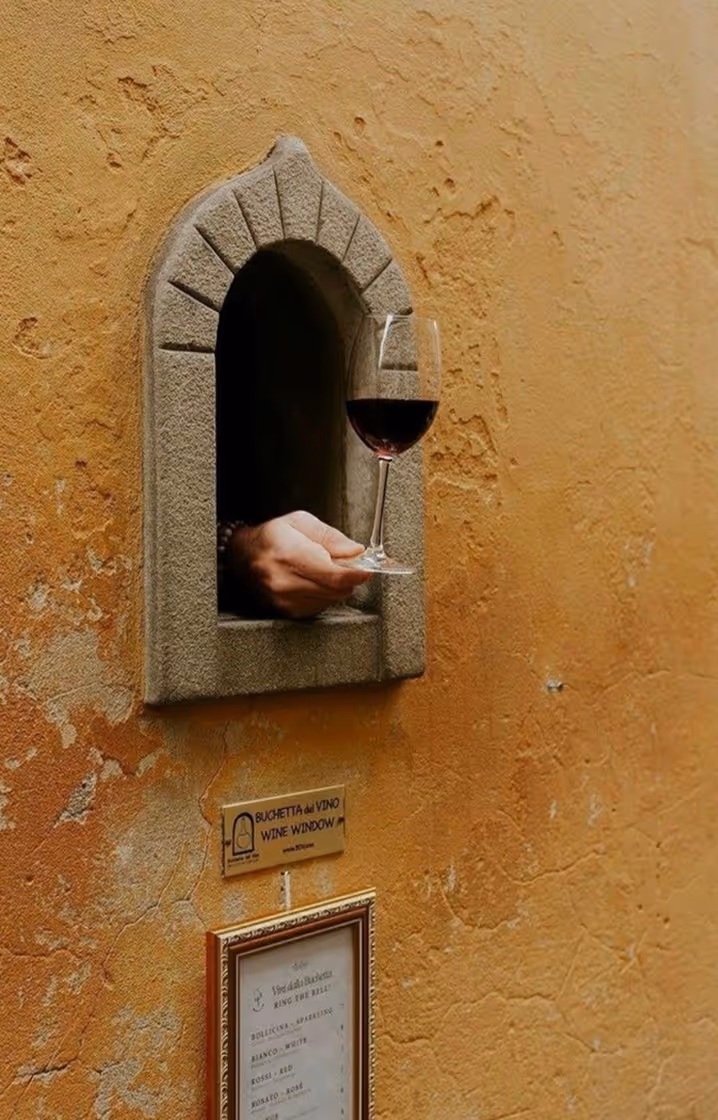 Рука, що простягає келих червоного вина через невелике аркове вікно у старовинній стіні з табличкою Wine Window. Follonica, Toscana