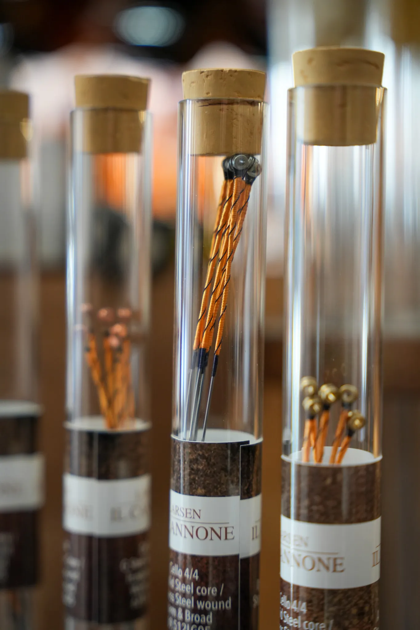 Cinq tubes en verre avec bouchons en liège contenant des cordes d'instruments de musique enveloppées d'un fil orange et noir.