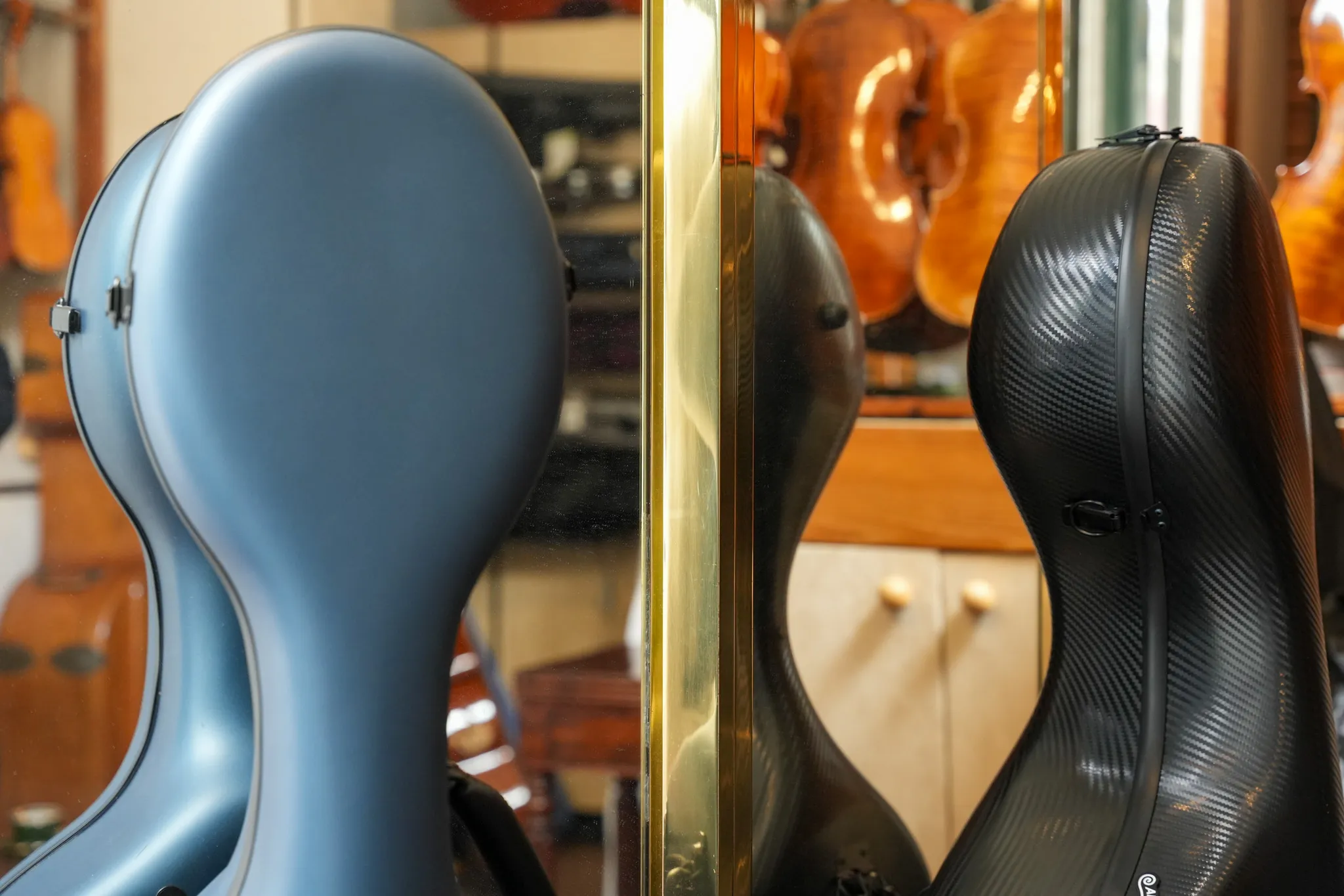 Deux étuis à violoncelle, l'un bleu mat et l'autre noir avec texture carbone, reflétés dans un miroir, avec des violoncelles en arrière-plan.