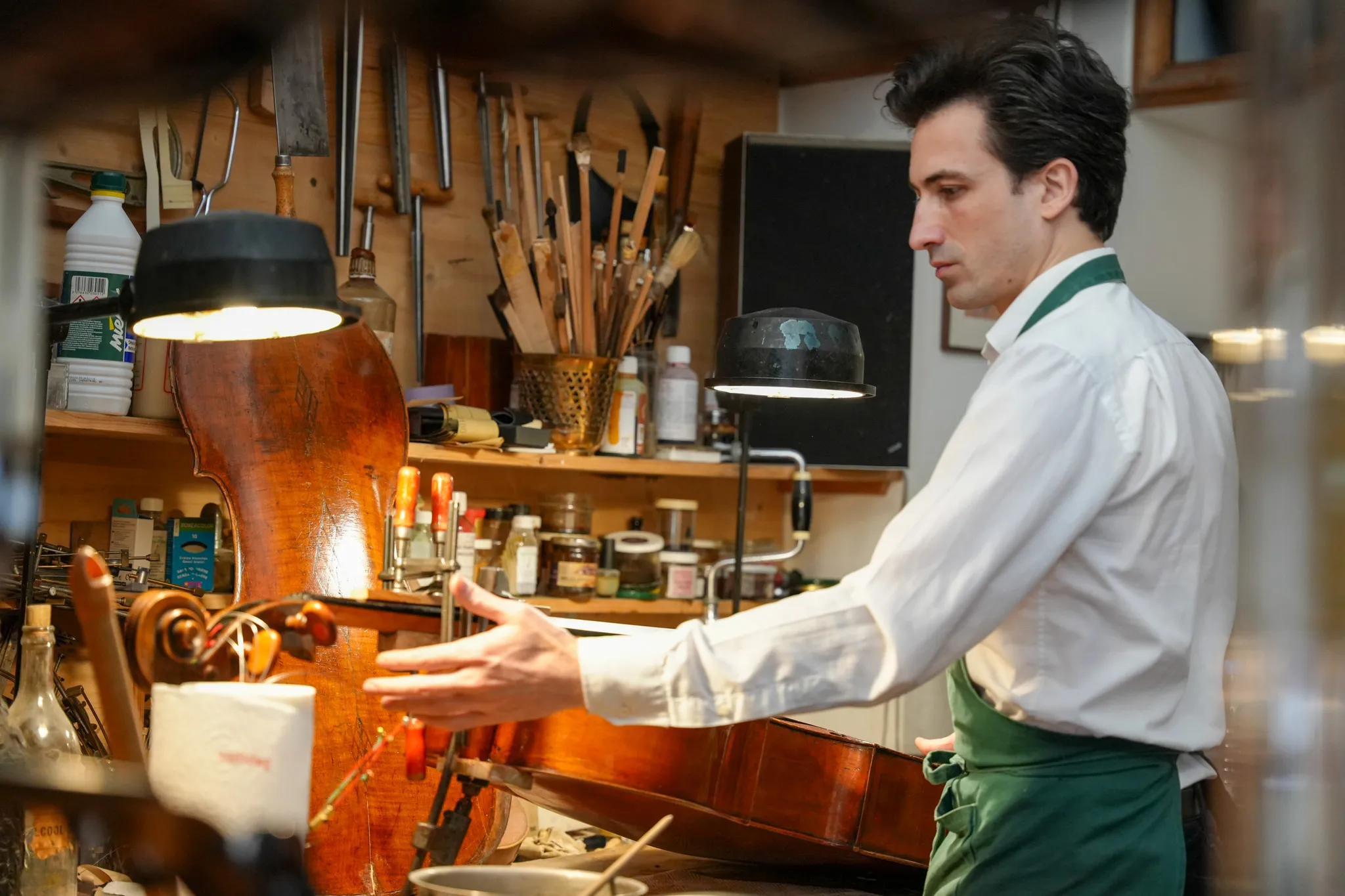 Un luthier ajustant un violoncelle dans son atelier avec divers outils et pinceaux en arrière-plan.