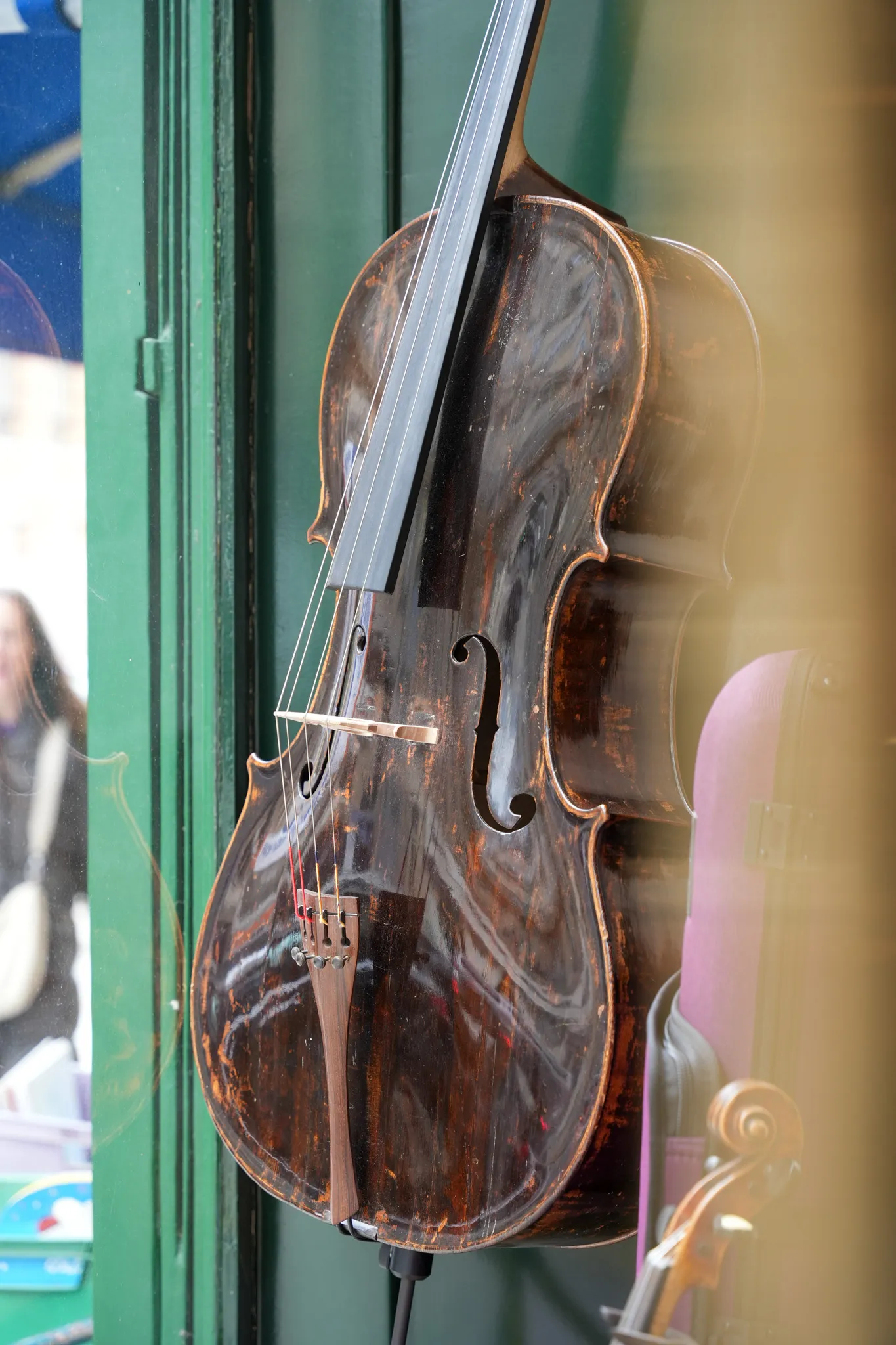 Un violoncelle ancien avec un vernis usé exposé devant une porte verte, avec un étui violet et un violon à côté.