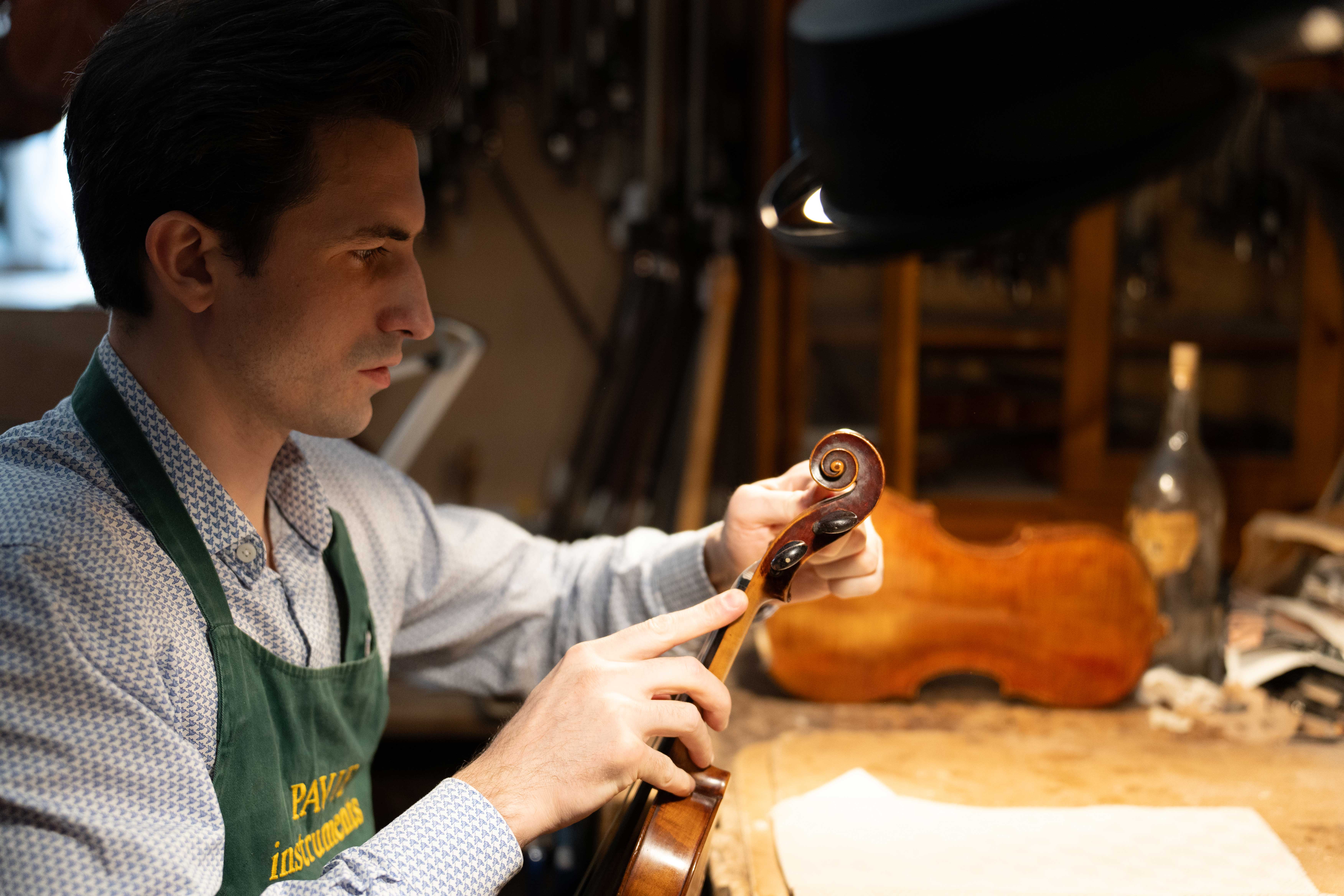 Un homme en tablier vert ajuste les cordes d'un violon dans un atelier de luthier.