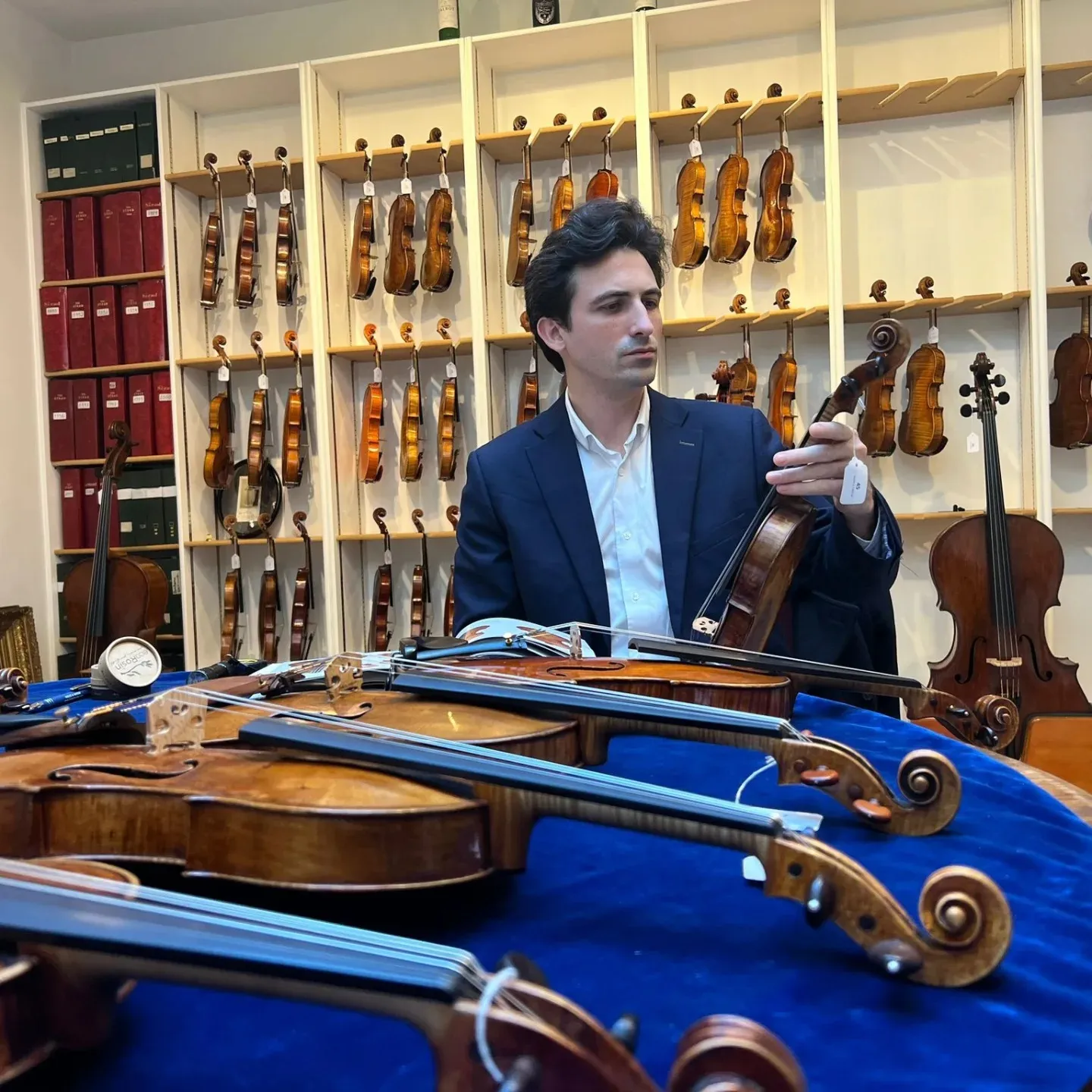 Un homme en costume bleu examine un violon dans un magasin rempli de violons suspendus et posés sur une table bleue.