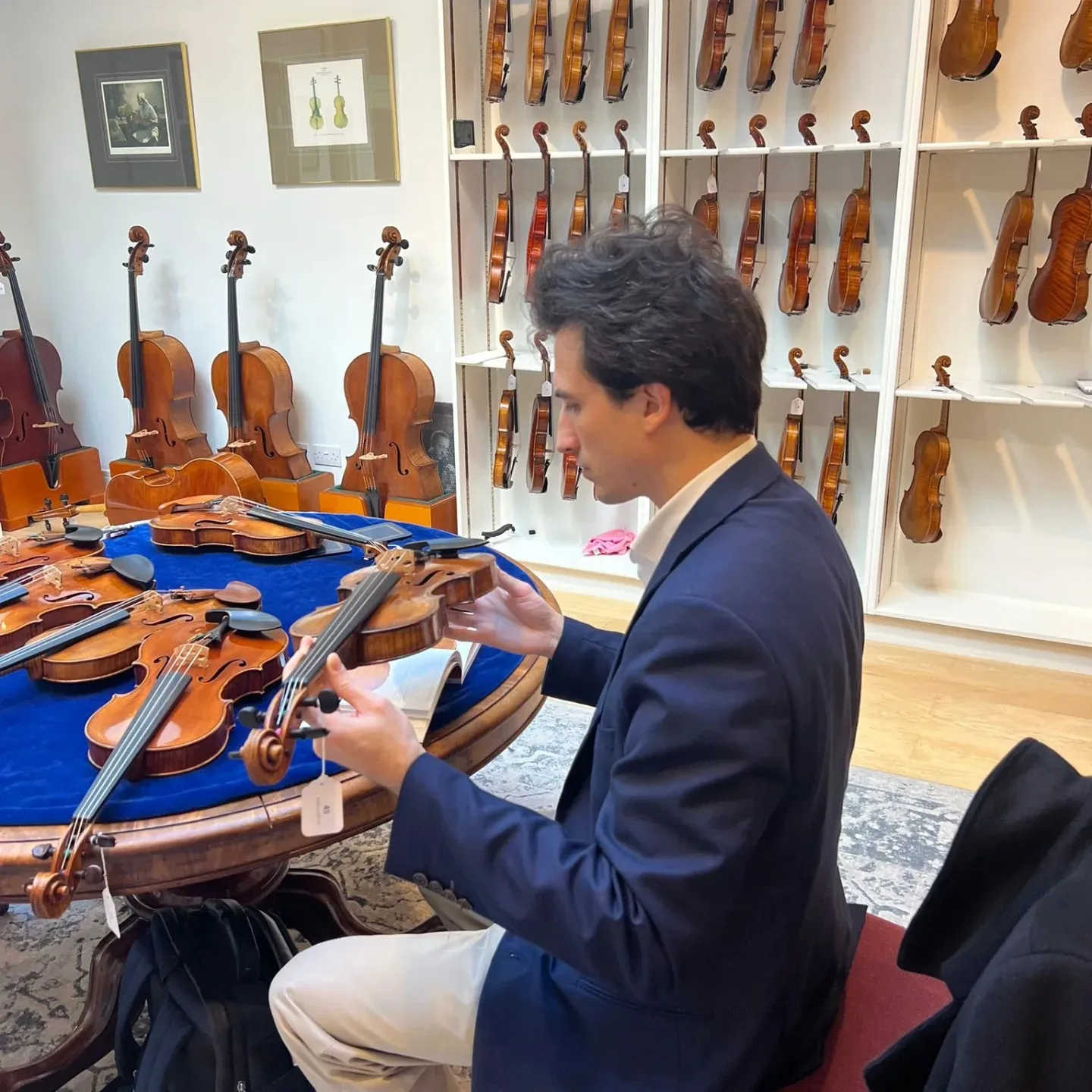 Un homme en veston bleu examine un violon assis à une table ronde couverte d'autres violons dans un magasin spécialisé.