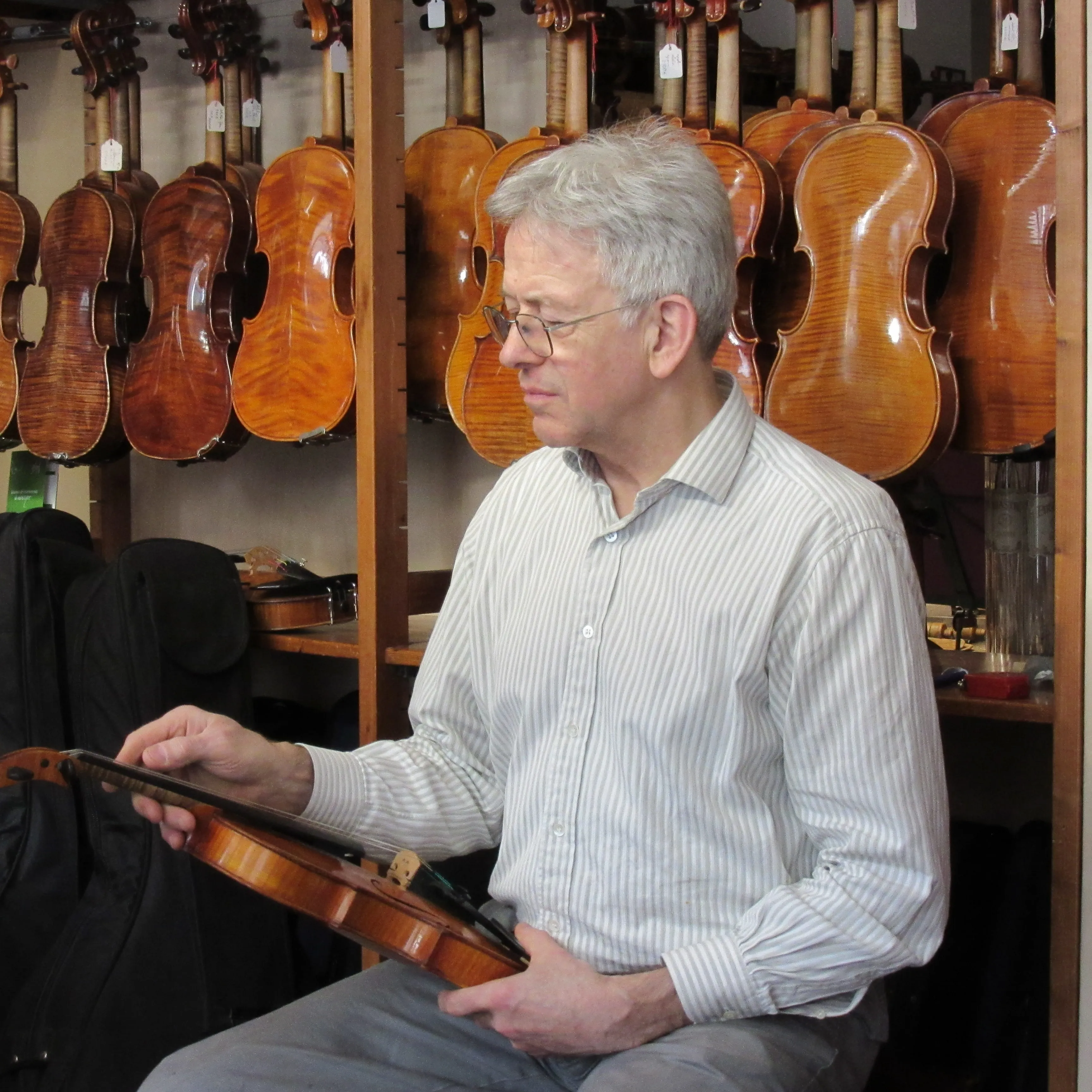 Un homme avec des cheveux gris et des lunettes tenant un violon dans une boutique avec plusieurs violons accrochés en arrière-plan.