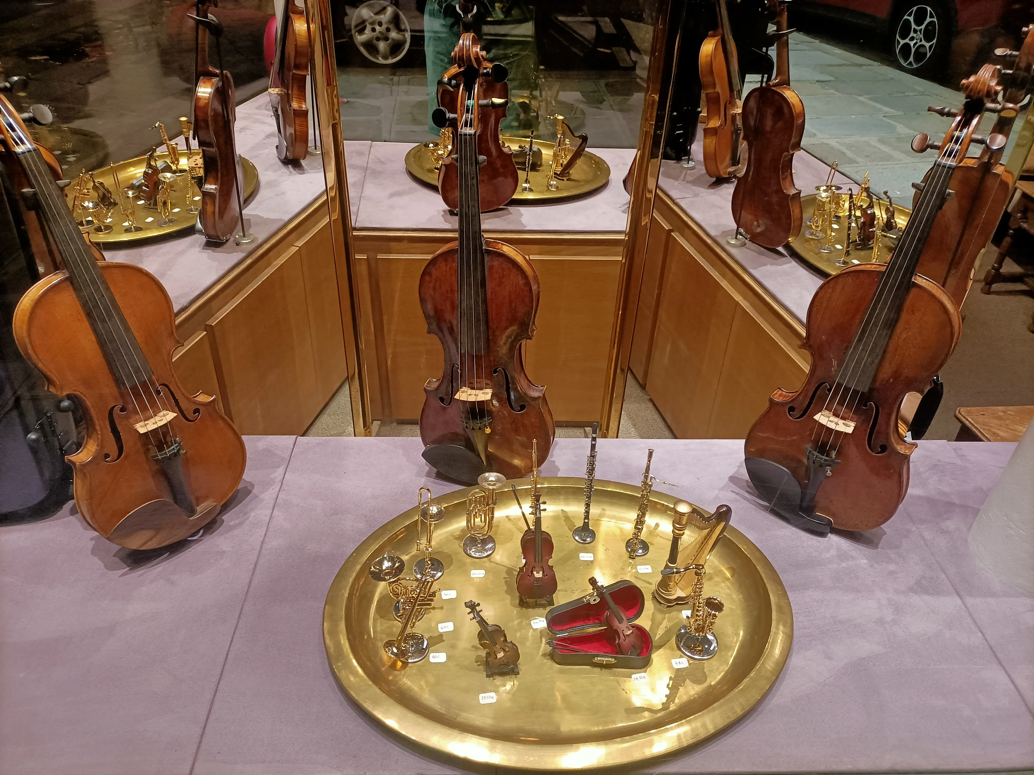Vitrine d'instruments de musique miniatures avec violons, trompettes, flûtes, harpe et saxophone sur un plateau doré, reflétés dans des miroirs.