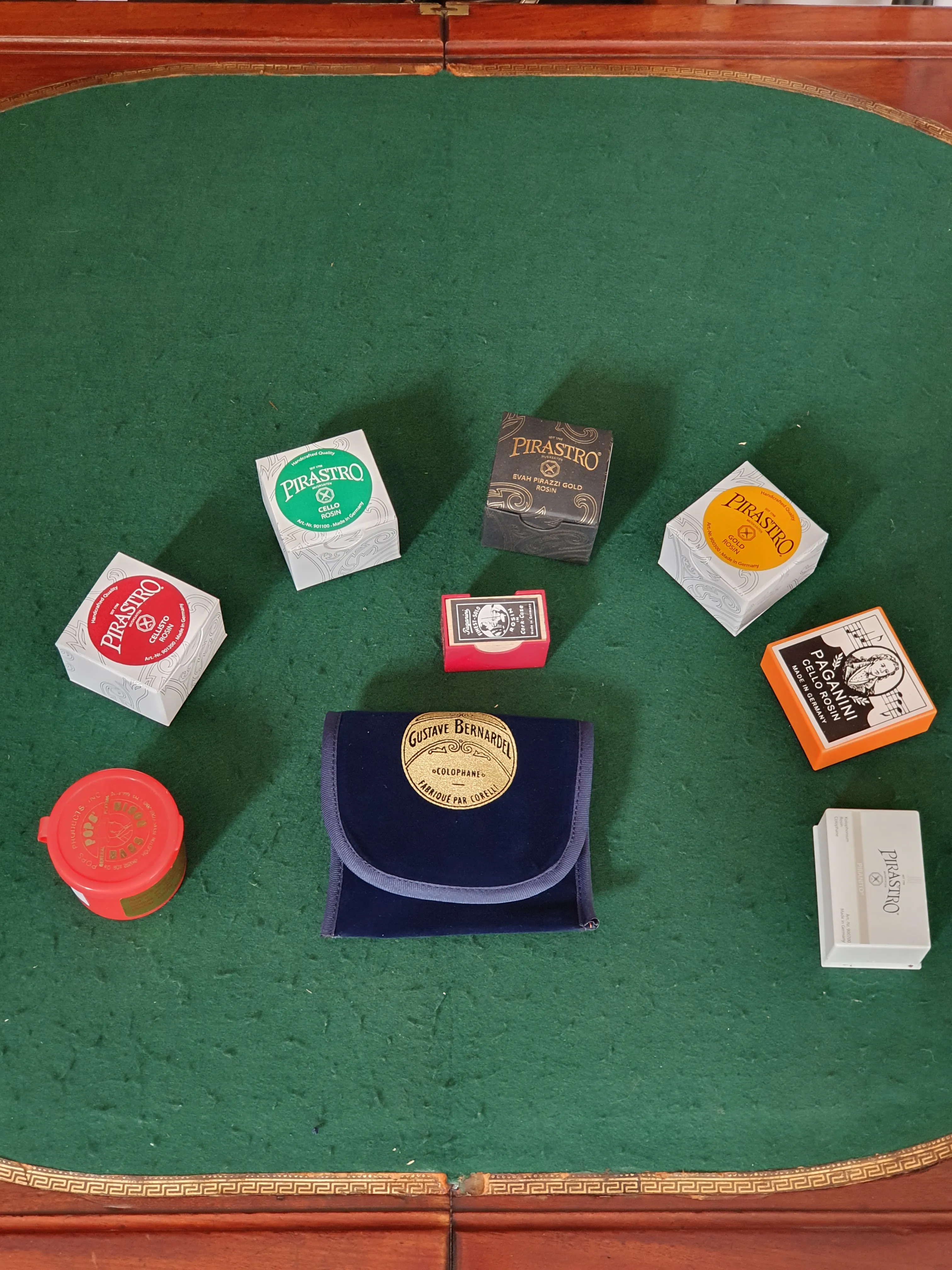 Plusieurs boîtes de colophane pour instruments à cordes disposées en cercle sur une surface verte, avec une pochette bleu foncé au centre.