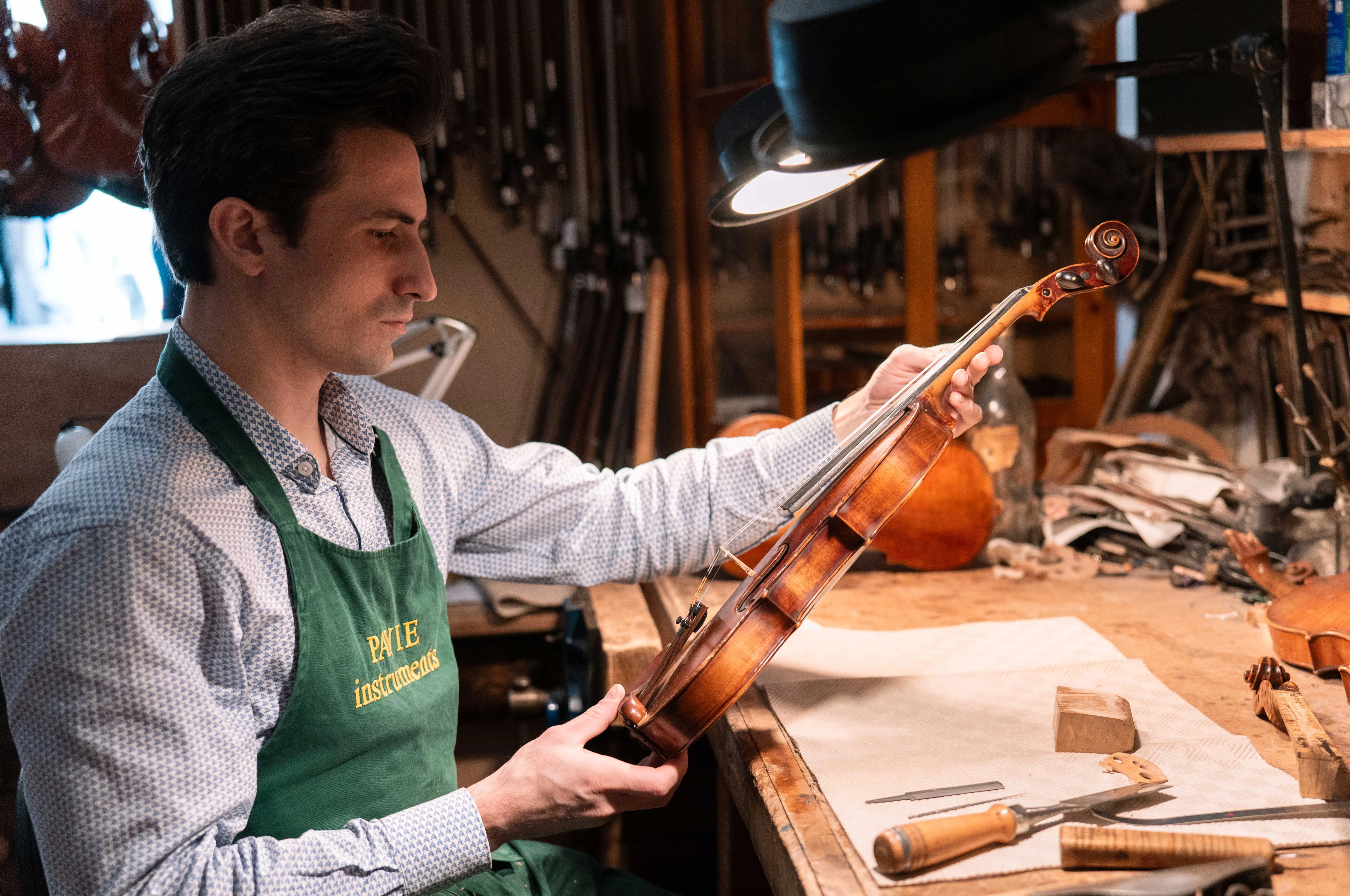 Luthier masculin examinant un violon dans un atelier rempli d'outils et d'instruments de musique.