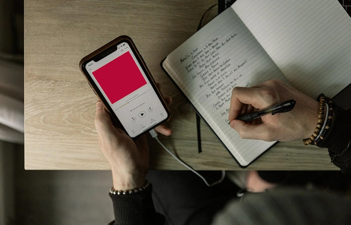 Person holding a smartphone playing audio while writing notes in a notebook on a wooden table.