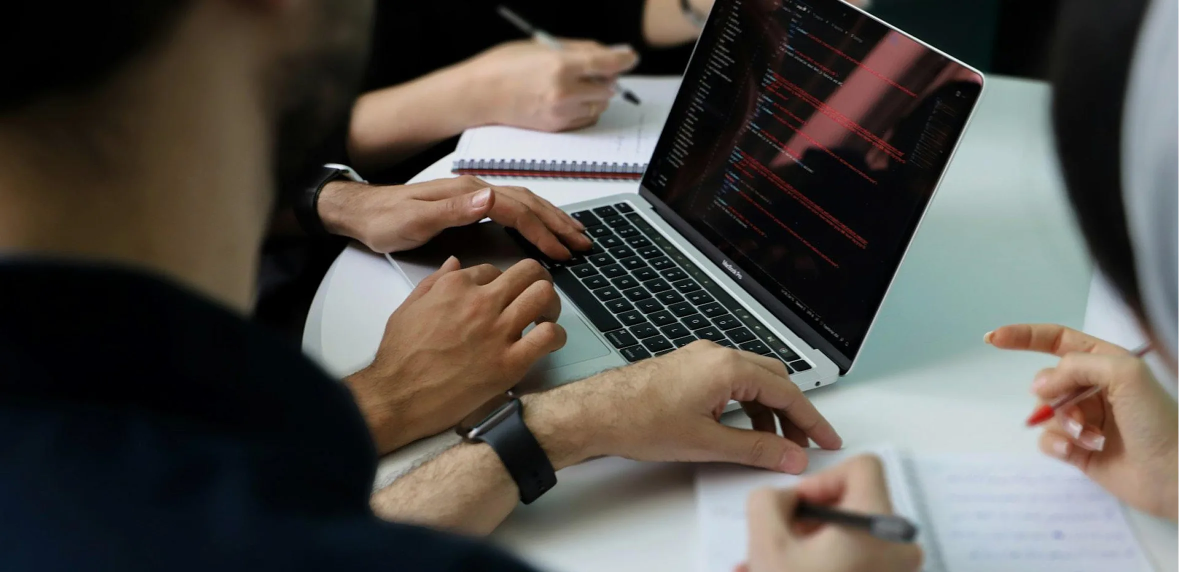 Two people working together on a laptop showing red code on a black screen, with others writing notes nearby.