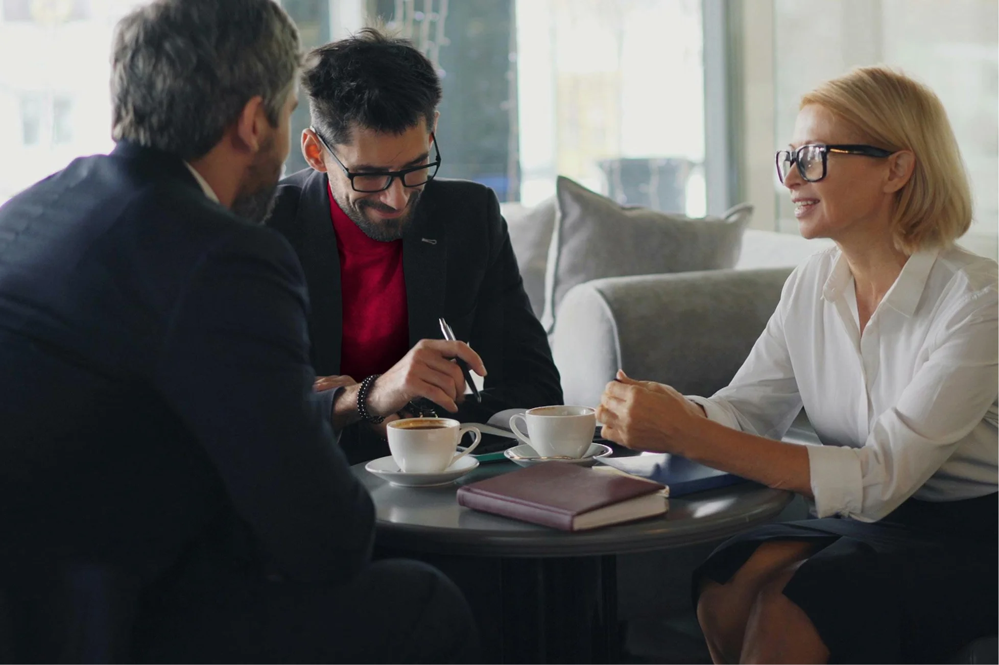 Three professionals in business attire having a discussion around a table with coffee cups and notebooks.