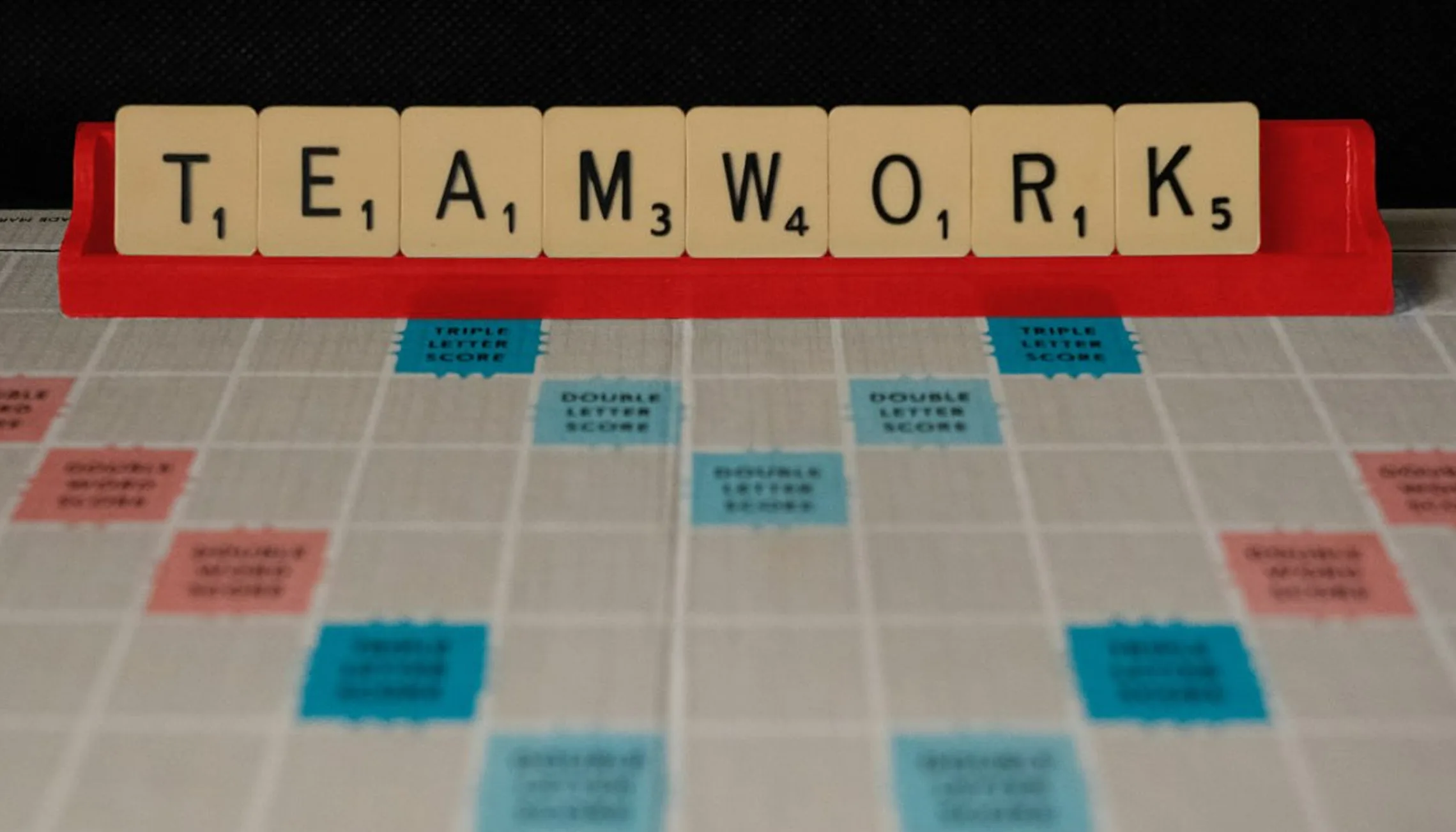 Scrabble tiles on a red rack spelling the word TEAMWORK above a Scrabble board.