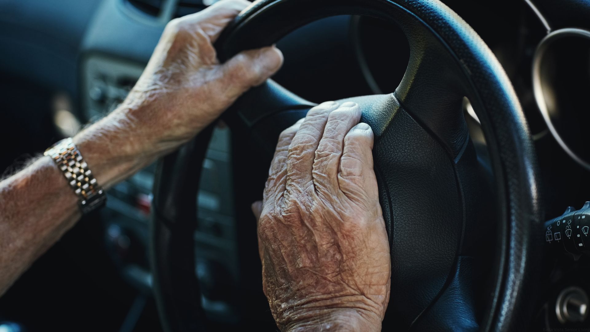 The hands of an older man rest on a car steering wheel.