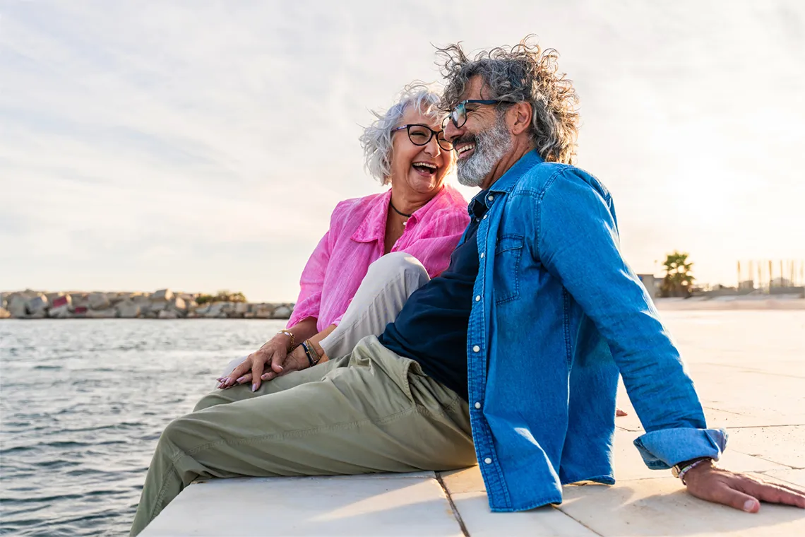 Older couple smiling and sitting by the water on a sunny day.