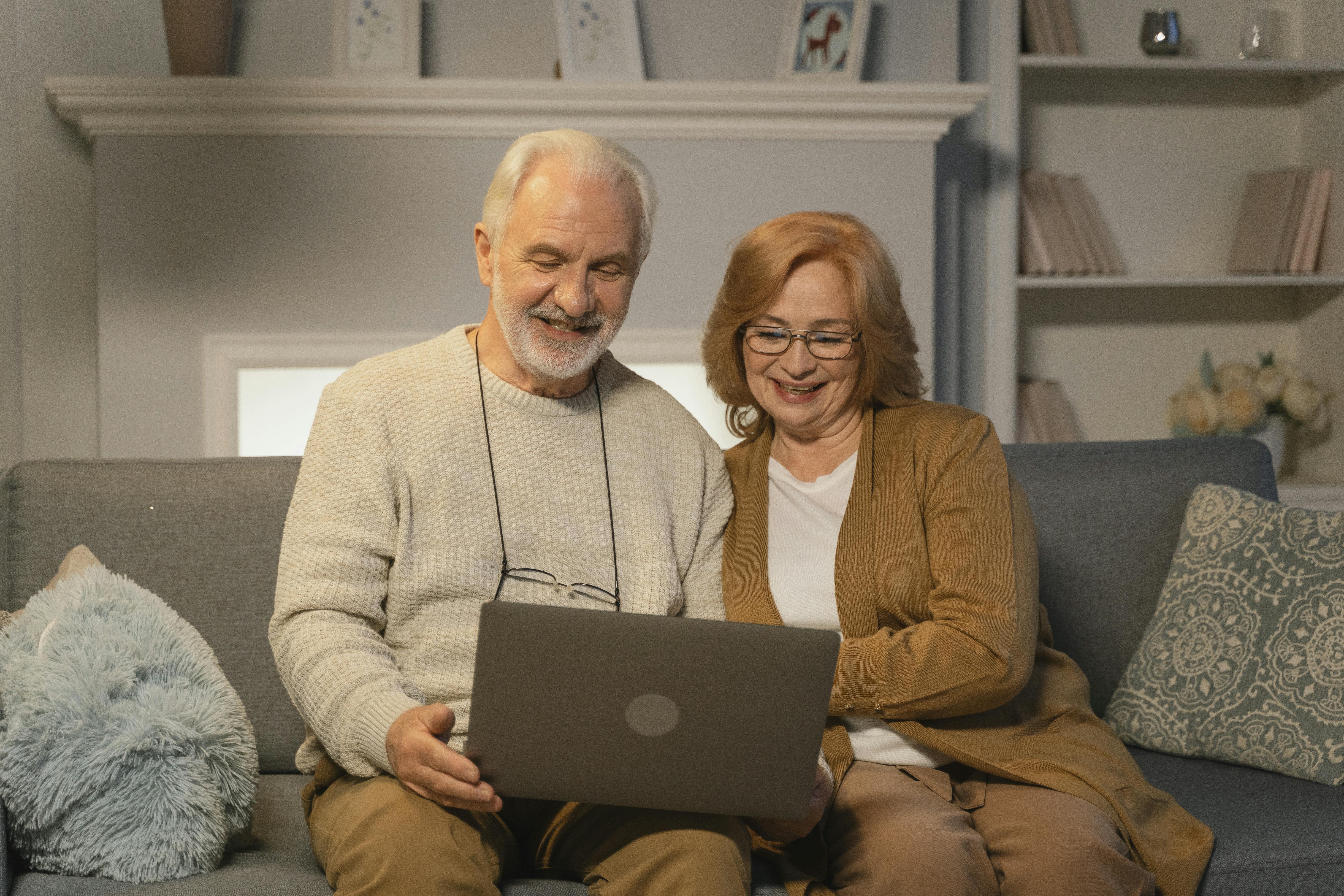 Smiling elderly couple sitting on a gray sofa and looking at a laptop together.
