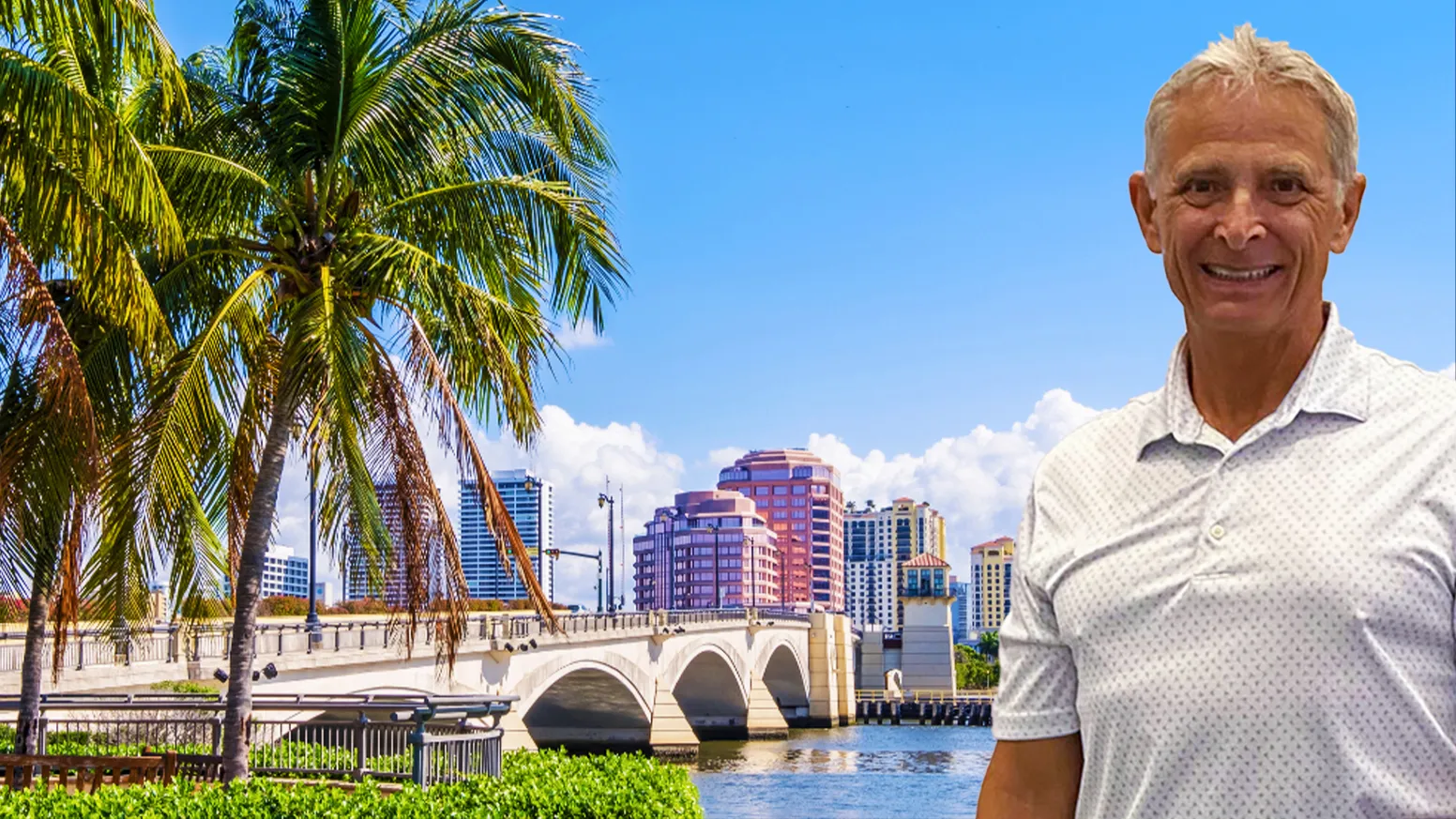 Garry Hiebert in a white polo shirt standing by West Palm Beach down town with palm trees, a bridge, and city buildings in the background under a blue sky.