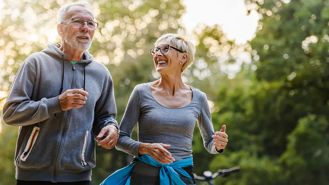 Smiling elderly couple jogging outdoors wearing glasses and casual athletic wear.