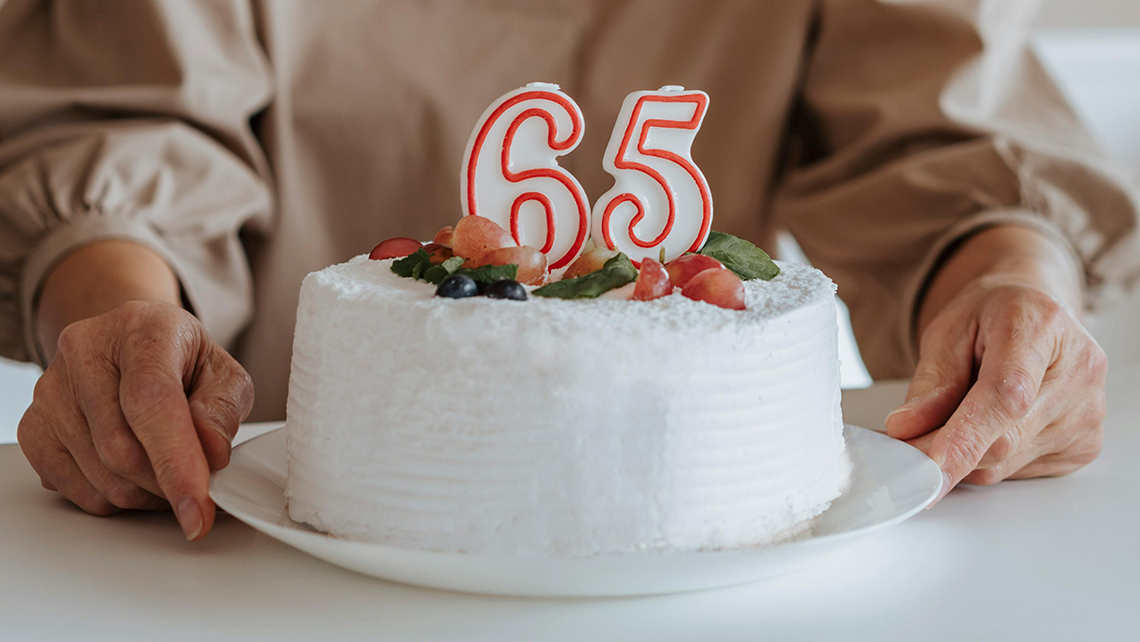 White frosted cake with fruit decorations and number candles 65 on top, held by an elderly person.