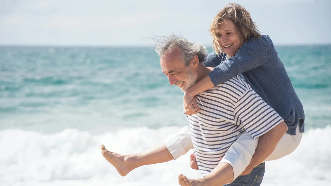 Smiling middle-aged man giving a piggyback ride to a woman by the ocean shore.