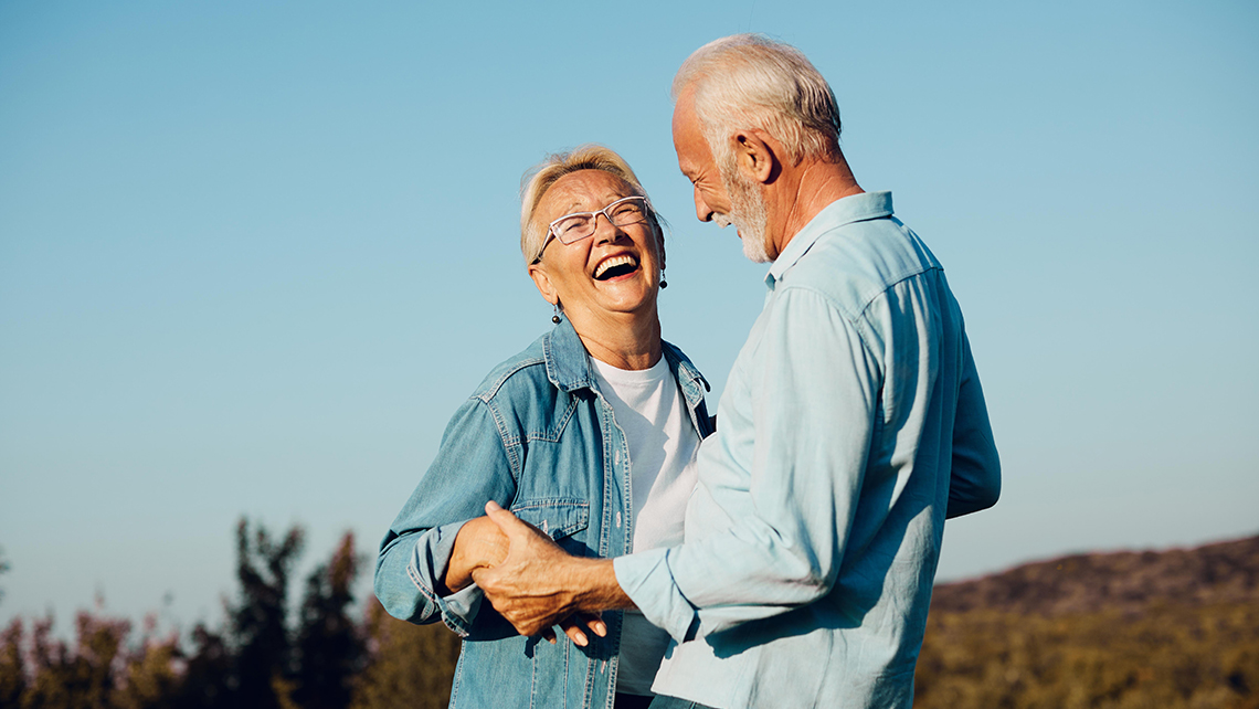Smiling elderly couple holding hands outdoors against a clear blue sky.