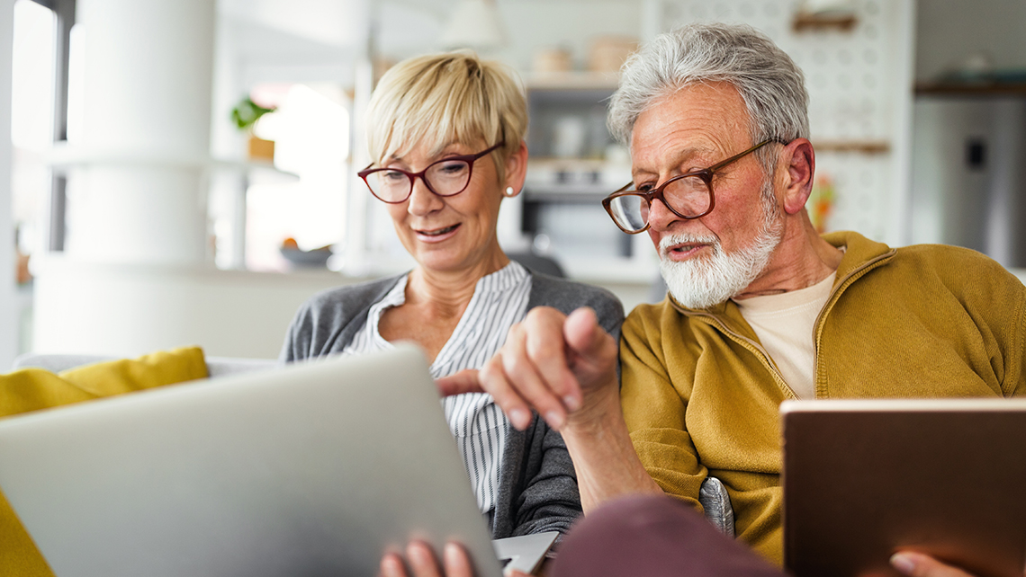Senior couple sitting on a couch looking at a laptop screen together in a bright living room.