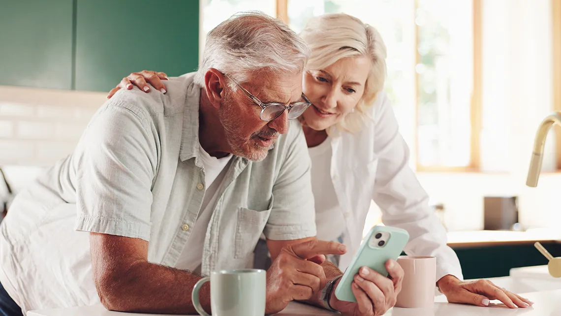 Senior couple leaning on kitchen counter, looking at a smartphone together with coffee mugs nearby.