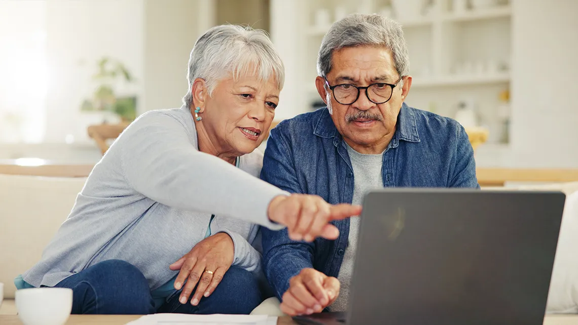 Senior couple sitting on a couch looking at a laptop, the woman pointing at the screen.