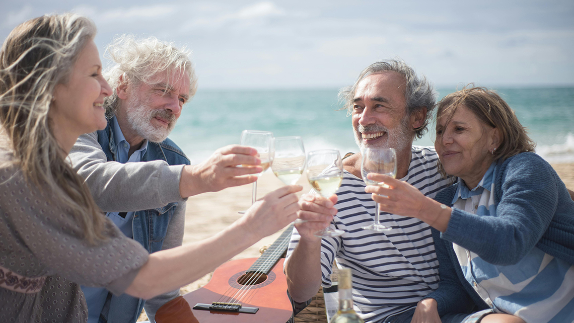 Four older adults sitting on a beach with a guitar, smiling and clinking wine glasses in a toast.