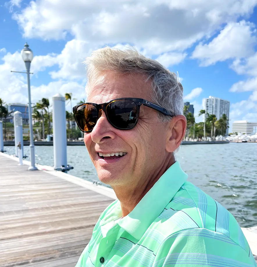 Four older adults sitting on a beach with a guitar, smiling and clinking wine glasses in a toast.