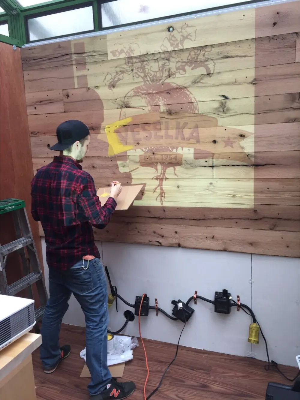 Man in a red plaid shirt and black cap painting a logo with the word 'VESELKA' on a wooden wall inside a room.