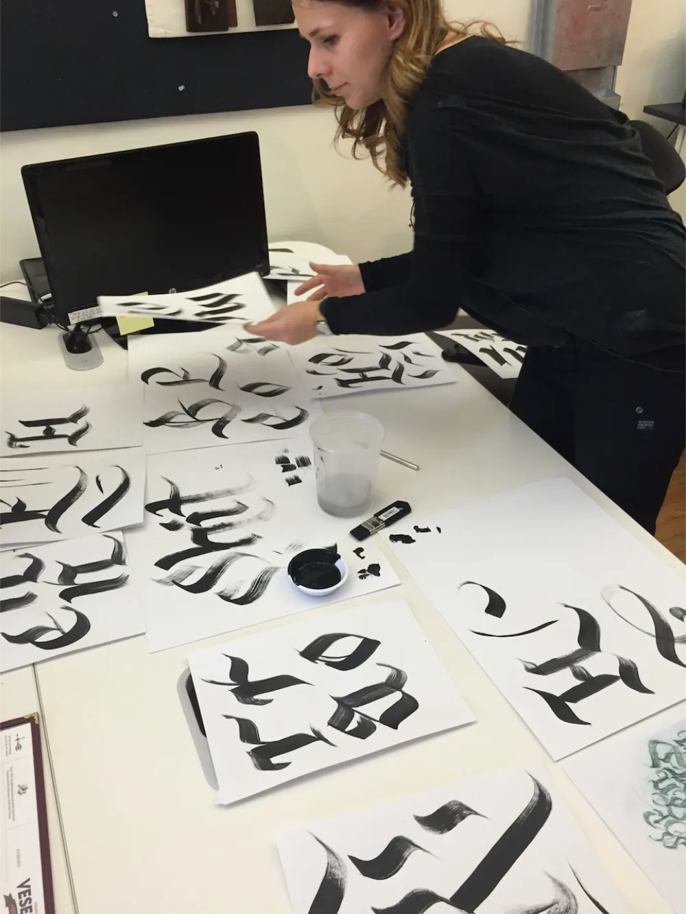 Person arranging sheets of paper with black calligraphy brush strokes on a white table next to a computer monitor.