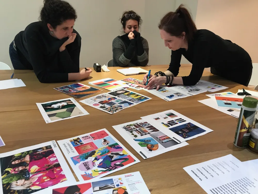 Three women leaning over a table covered with colorful printed materials and design samples, collaborating in a creative work session.