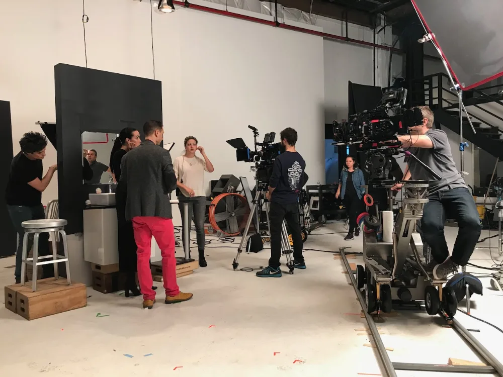 Film crew on a studio set with cameras on tripods and dolly tracks, preparing to shoot a scene with actors and crew members talking.