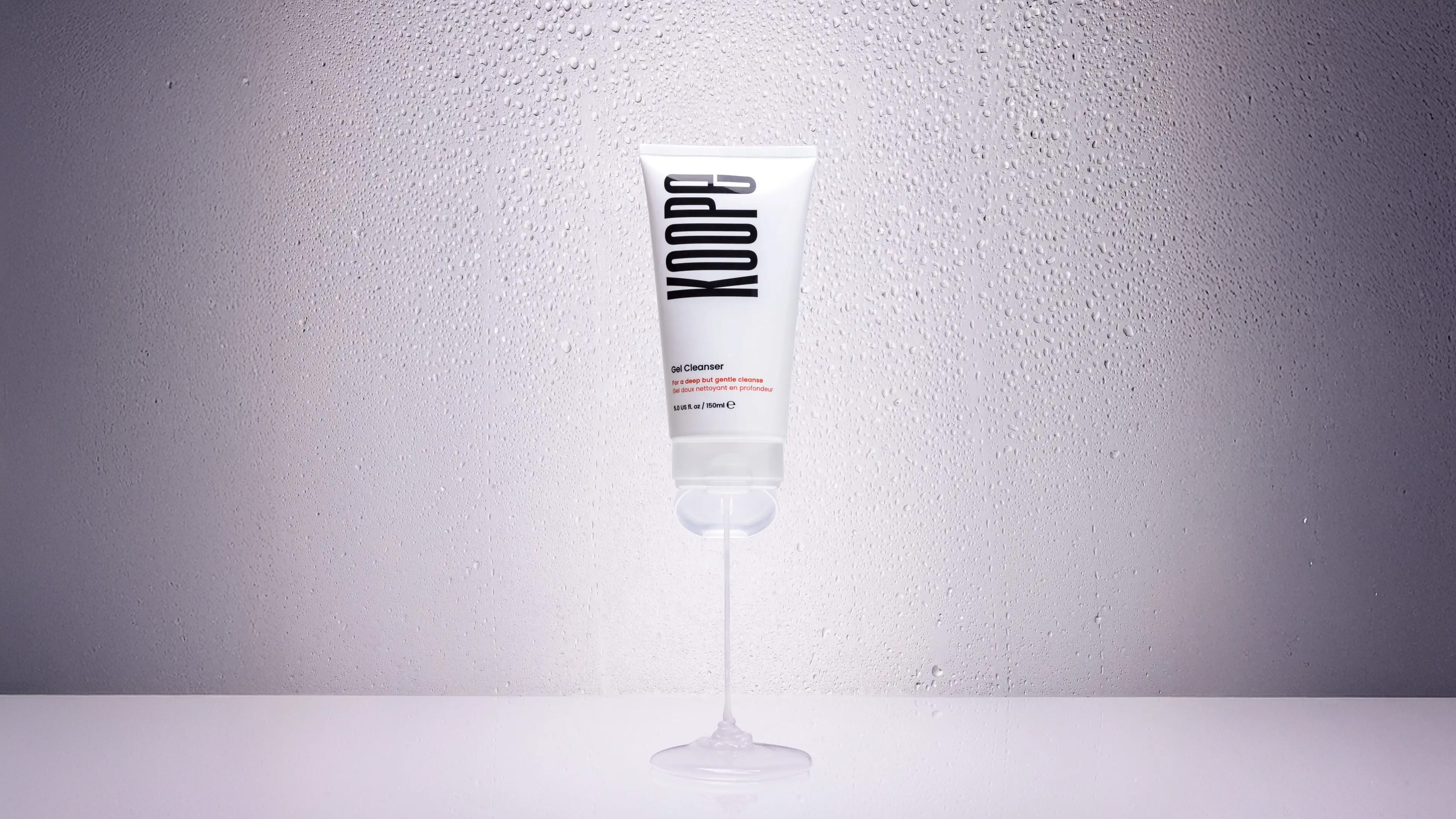 A white tube of KOOPe gel cleanser with liquid dispensed on a wet surface and a water-drop background.