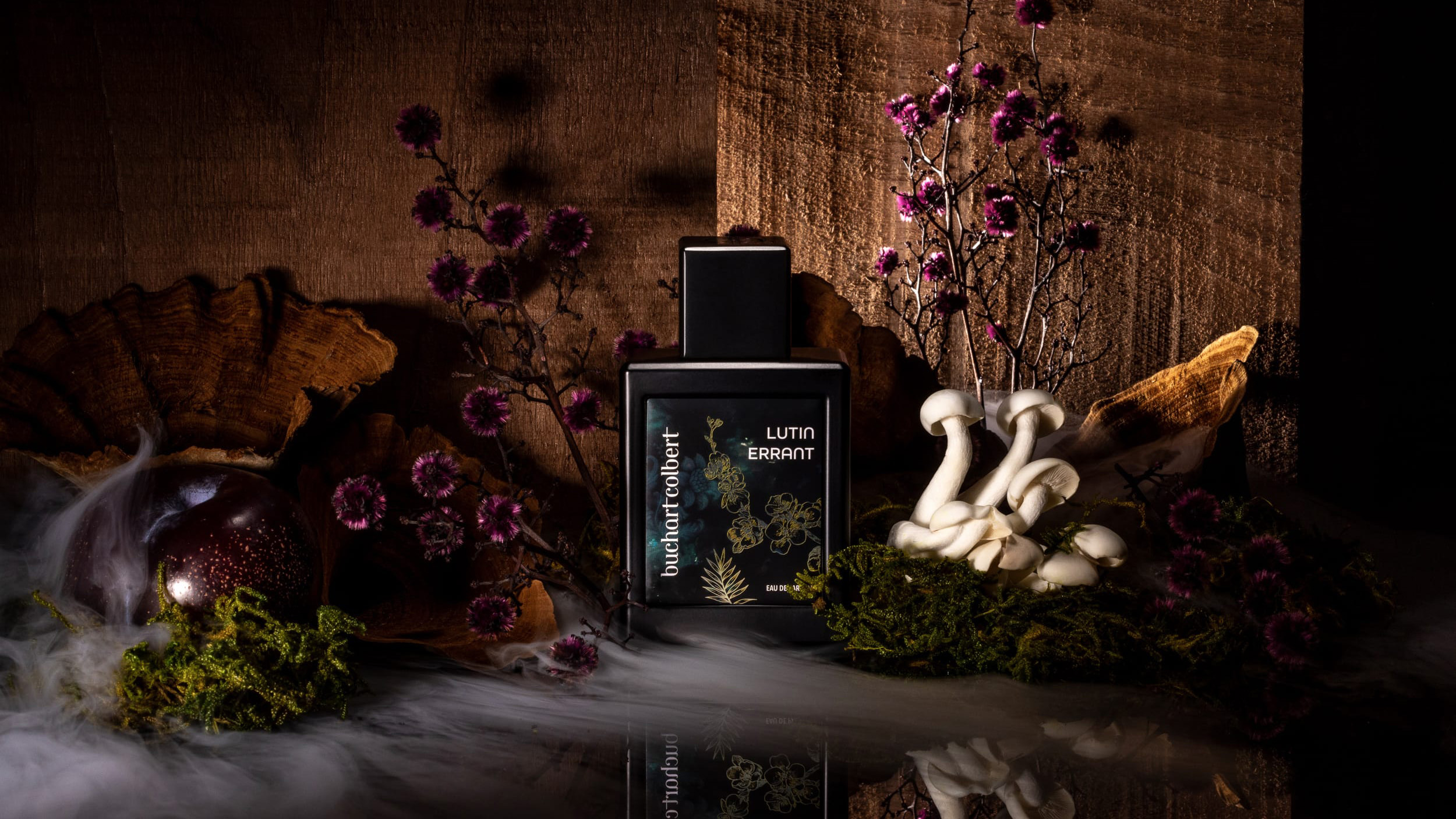 Black perfume bottle labeled Lutin Errant among moss, white mushrooms, purple flowers, and wood background.