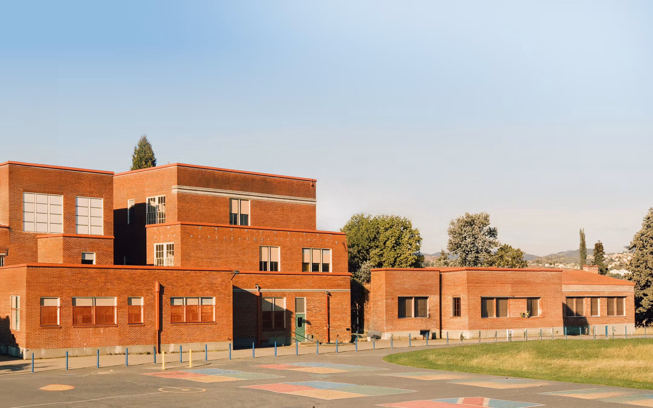 Red brick school building with multiple windows next to a playground with colorful markings.