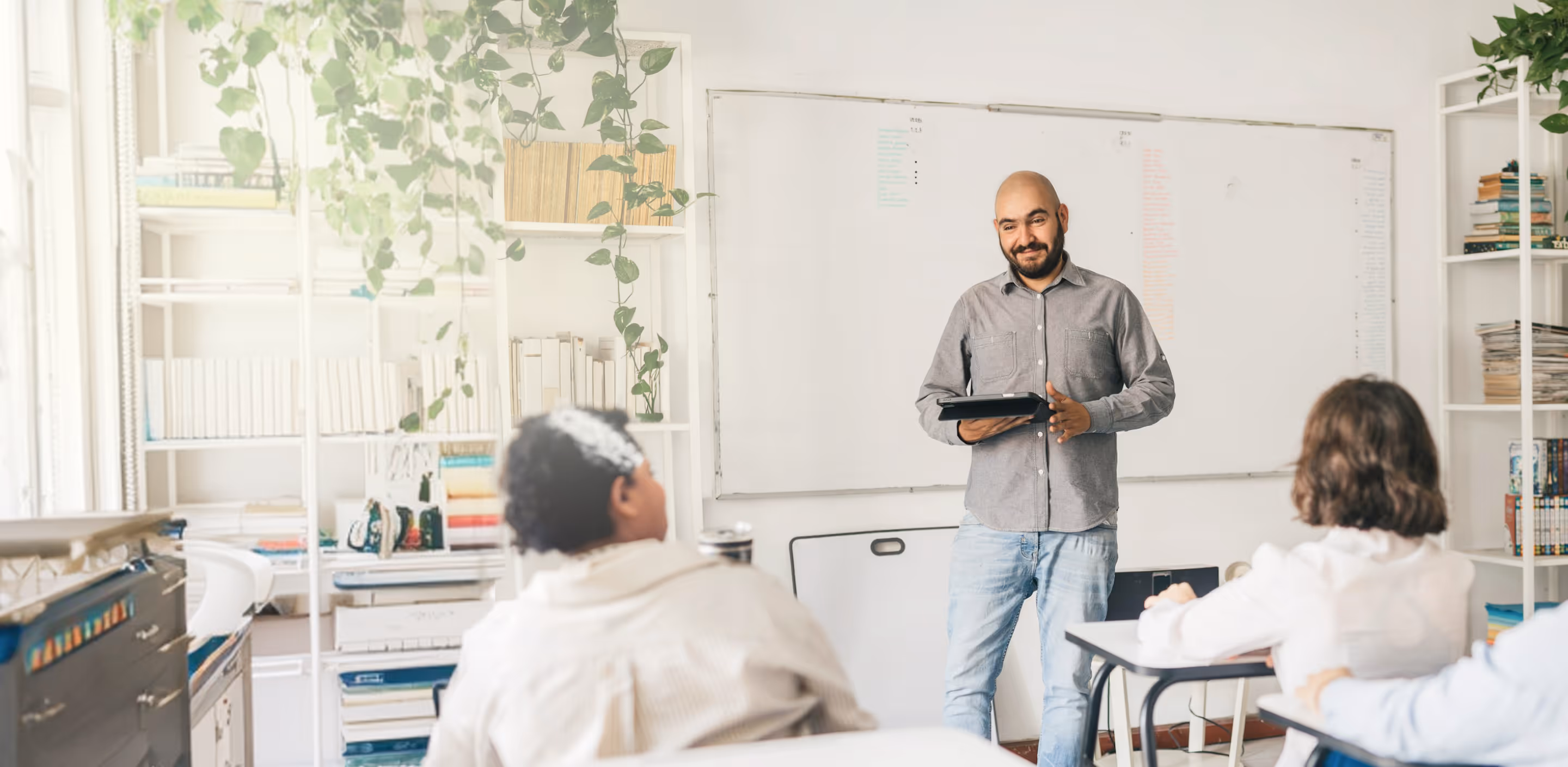 Teacher holding a tablet while standing in front of a whiteboard teaching students at desks.