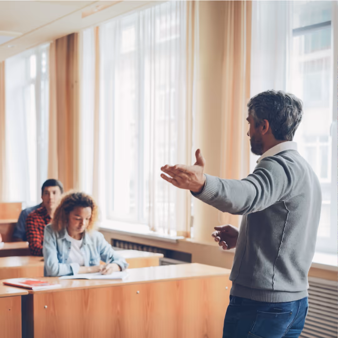 Teacher speaking to students seated at desks in a bright classroom with large windows.