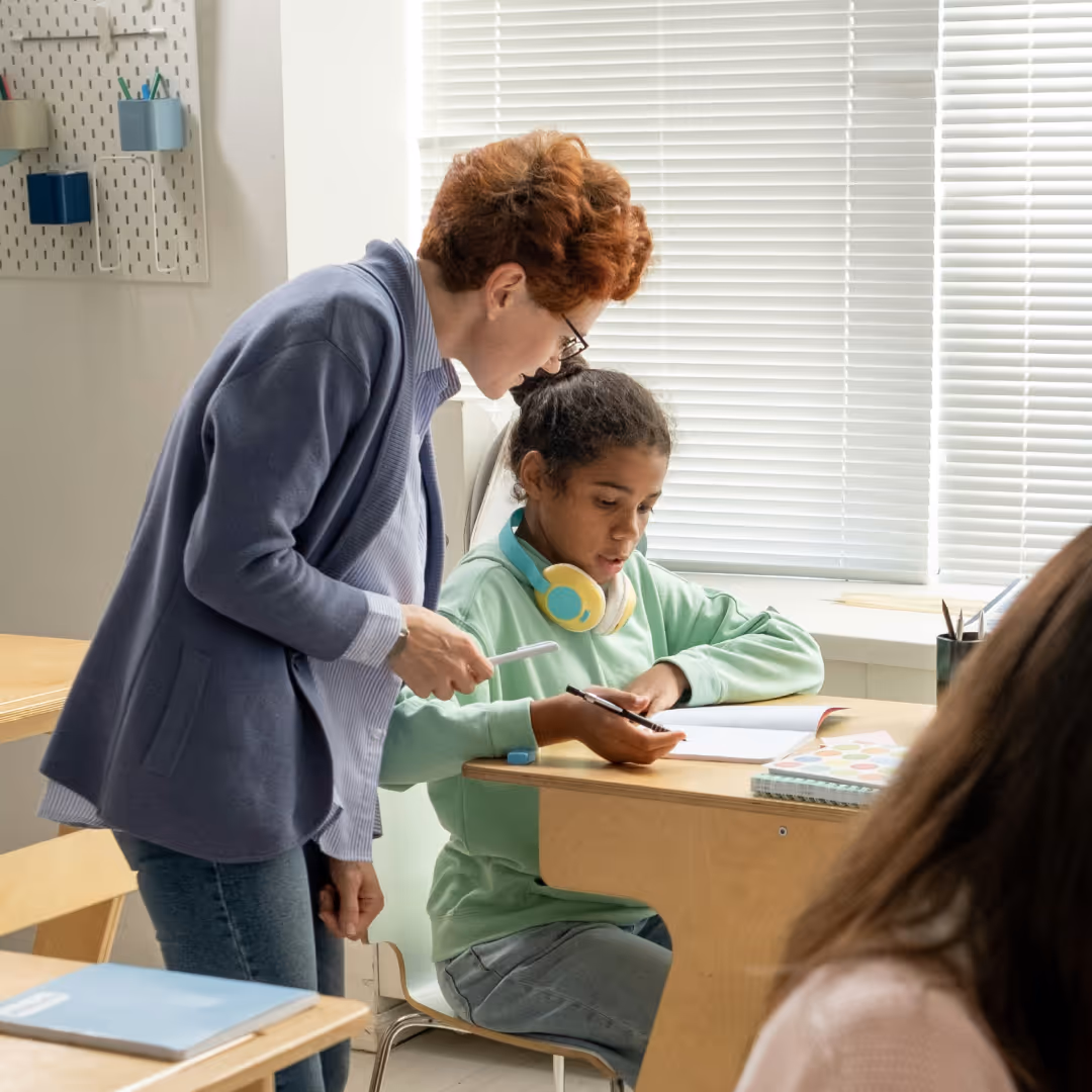 Teacher helping student with homework at desk in classroom with blinds on window.