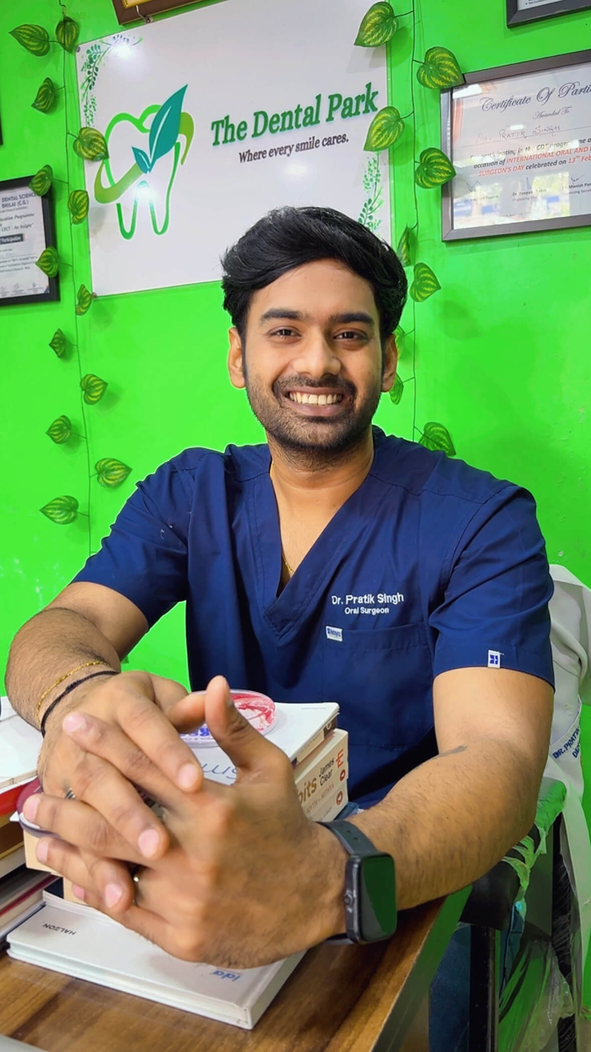 Smiling male oral surgeon in blue scrubs sitting at a desk with dental books and certificates on a bright green wall.